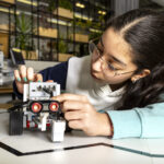 Ein 14-jähriges Mädchen in einem weißen Pullover mit dunkelblauen und hellblauen Ärmeln lehnt sich auf den Tisch und verkabelt den Roboter. Der Roboter mit schwarzen Rädern, weißen Bauteilen und zwei auffälligen roten Augen steht auf einer hellen Oberfläche mit einer schwarzen Linie, die einen Arbeitsbereich suggeriert. Die Nägel des Mädchens sind türkis lackiert. Das Bild ist waagerecht aufgenommen.
